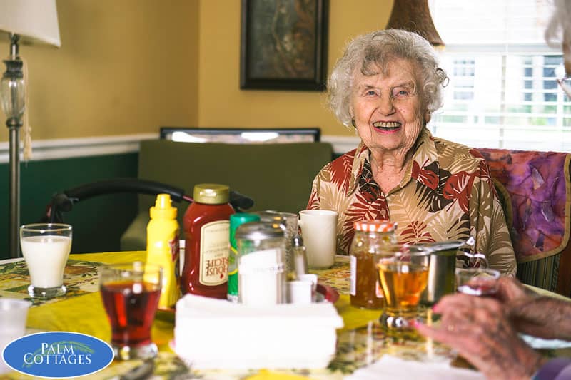elderly resident with several others (only hands pictured) around a crowded dining table enjoying breakfast and laughing
